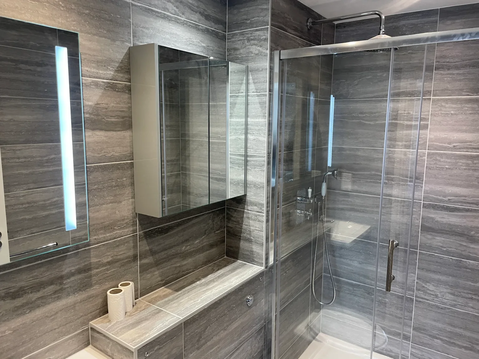 Completed compact client shower room with grey tiles, illuminated mirror and glass shower